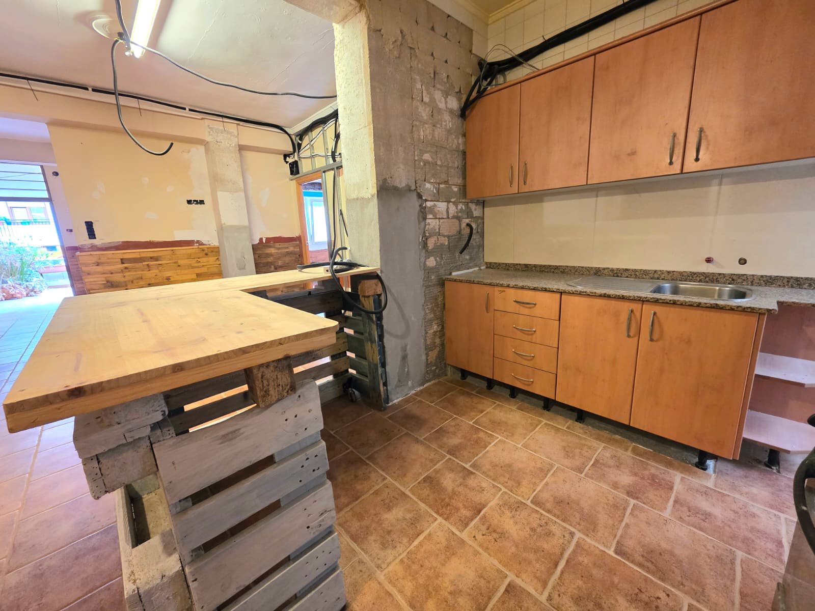 Espacio de cocina en reforma con mobiliario de madera, encimera de granito, suelo de gres rústico y una estructura de isla realizada con palés.