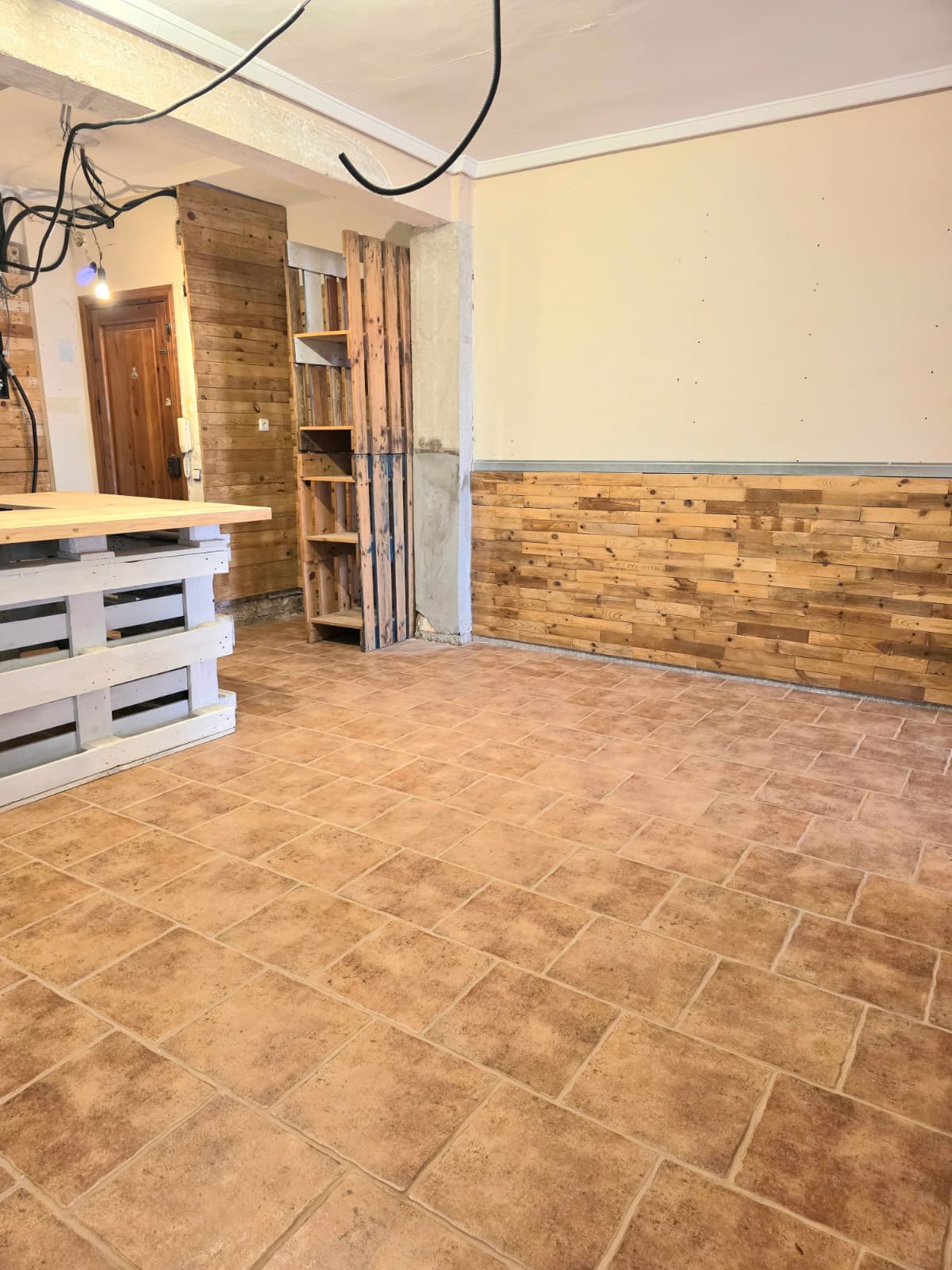 Espacio interior con suelo de baldosas cerámicas tipo terracota y friso de madera rústica. Incluye un mostrador de palets blancos y estanterías de madera natural.