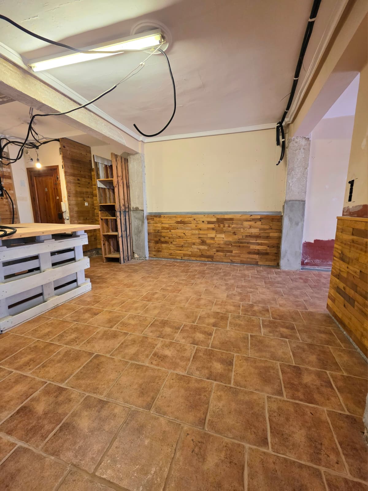 Espacio interior con suelo de baldosas tipo terracota, friso de madera rústica y un mostrador artesanal de palés blancos con encimera de madera clara.