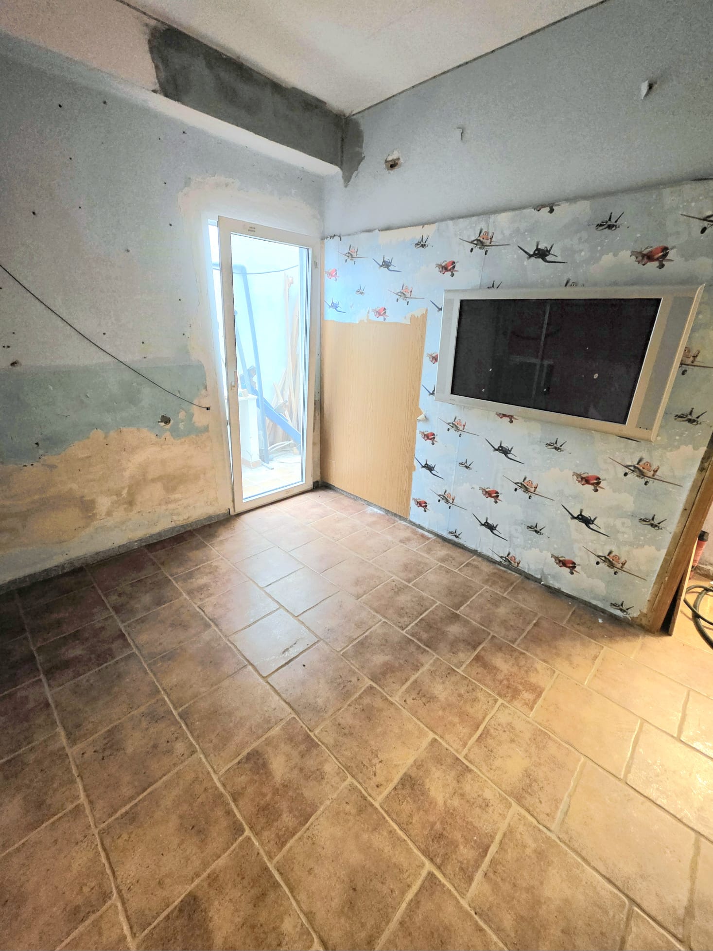 Espacio interior en reforma con suelos de gres cerámico, puerta de cristal con marco blanco y pared con papel pintado de aviones y soporte para pantalla integrado.