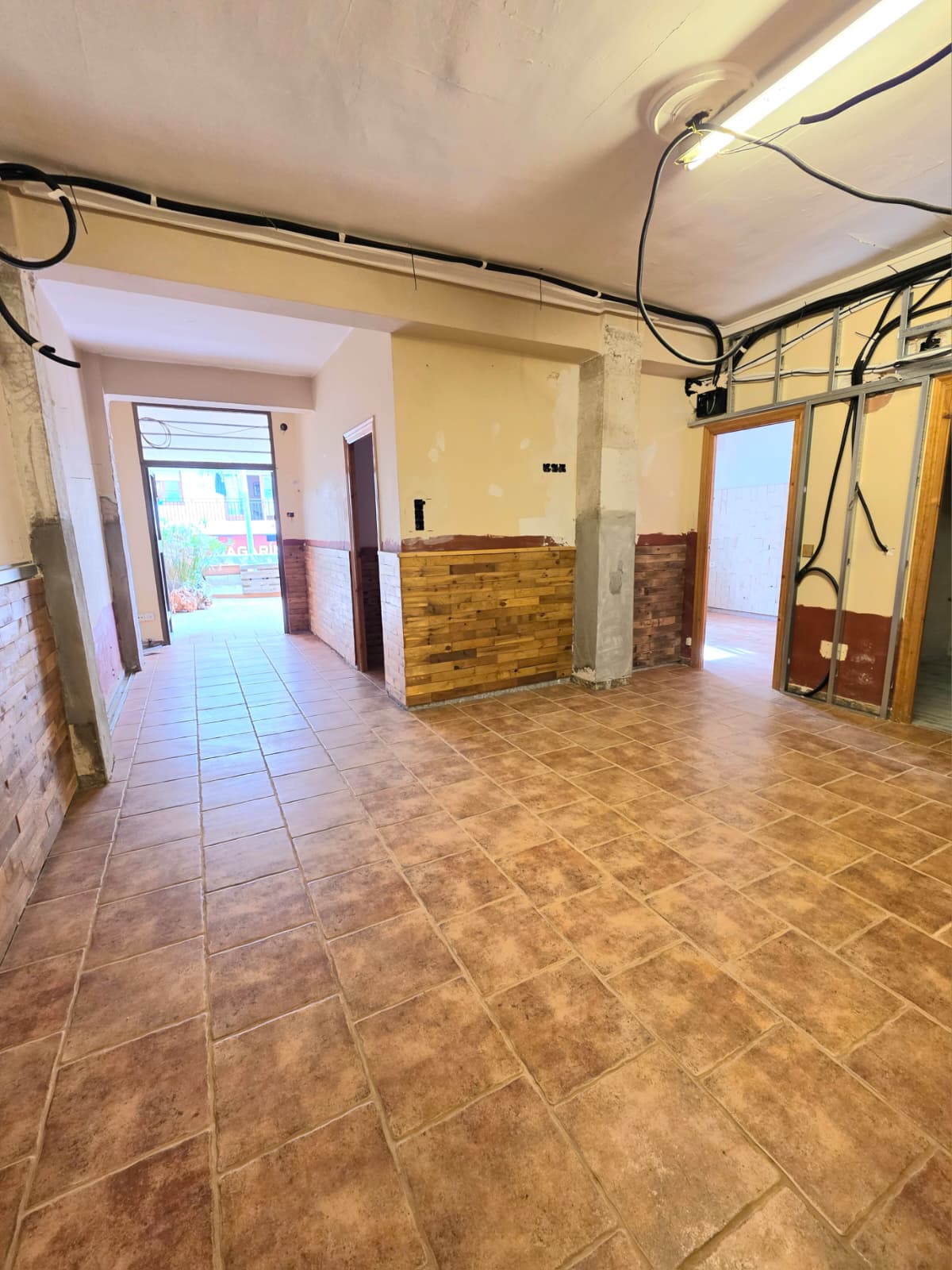 Espacio interior en reforma con suelos de gres rústico, friso de madera a media altura e instalaciones eléctricas y perfiles metálicos a la vista.