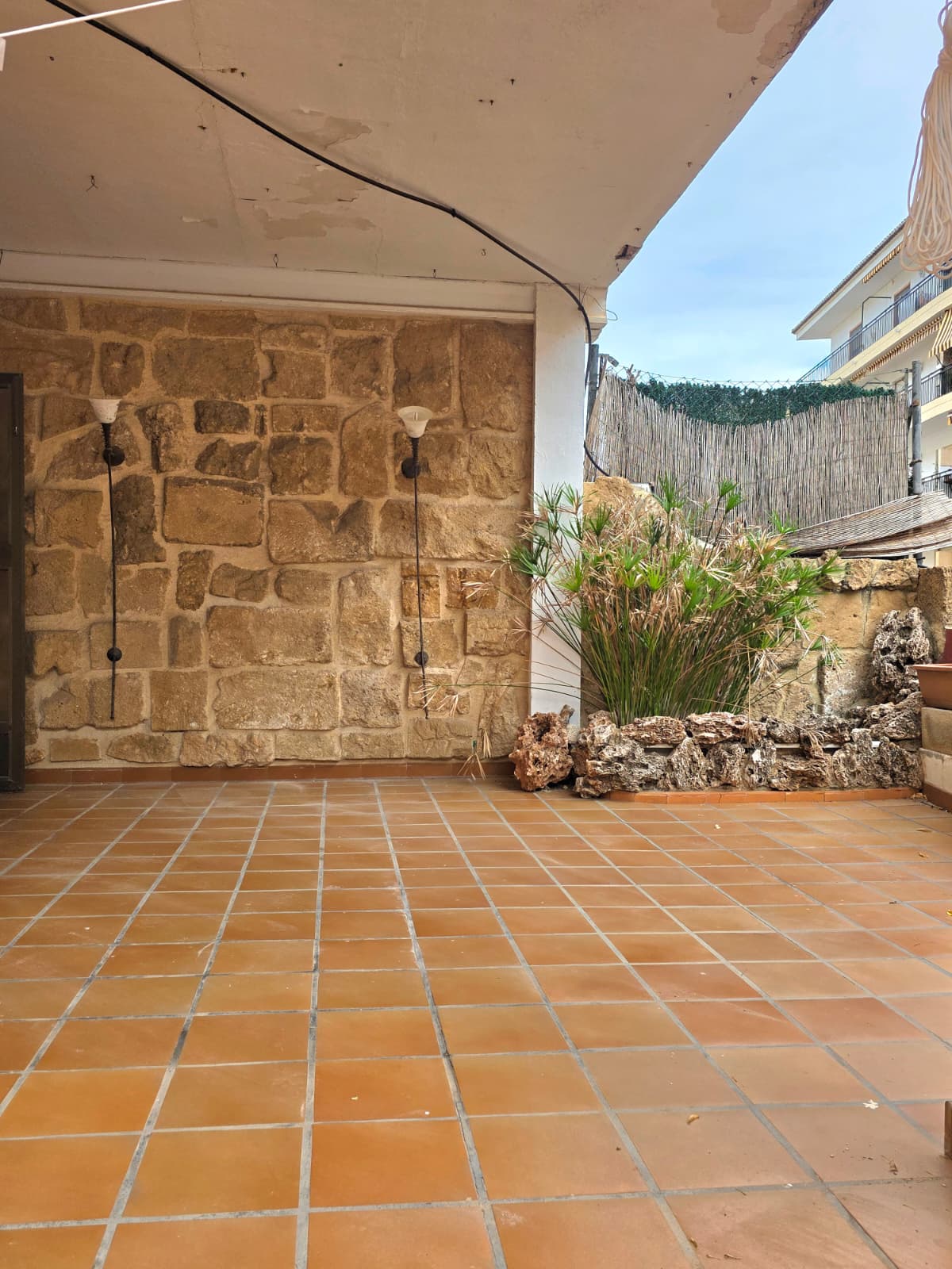 Terraza cubierta con suelo de baldosas de terracota, pared de piedra rústica con apliques de iluminación y jardinera decorativa con rocas naturales.