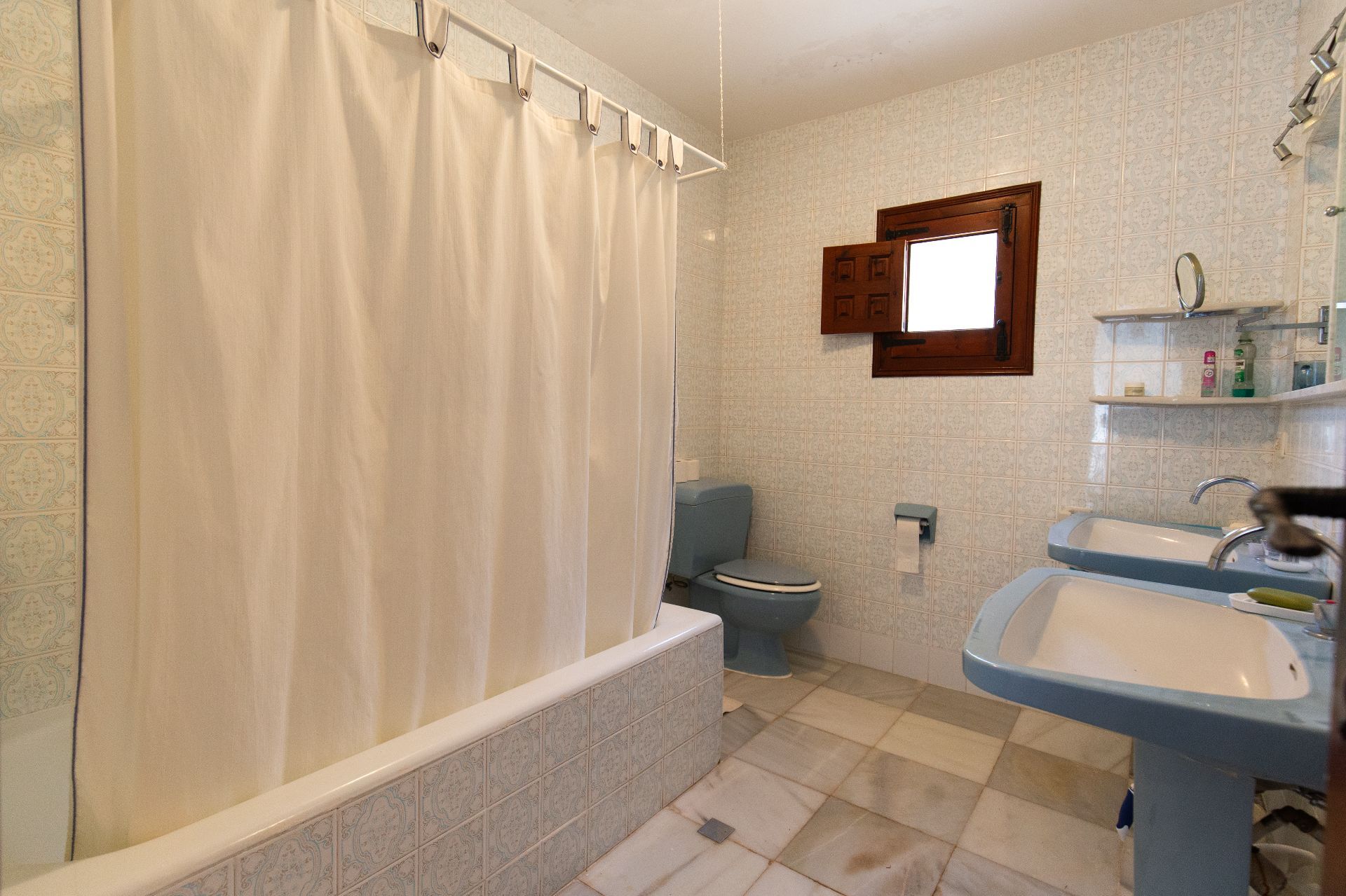 Baño de estilo tradicional con lavabos dobles de pedestal azul, inodoro a juego y bañera alicatada. Presenta azulejos decorados y una ventana de madera con contraventana.