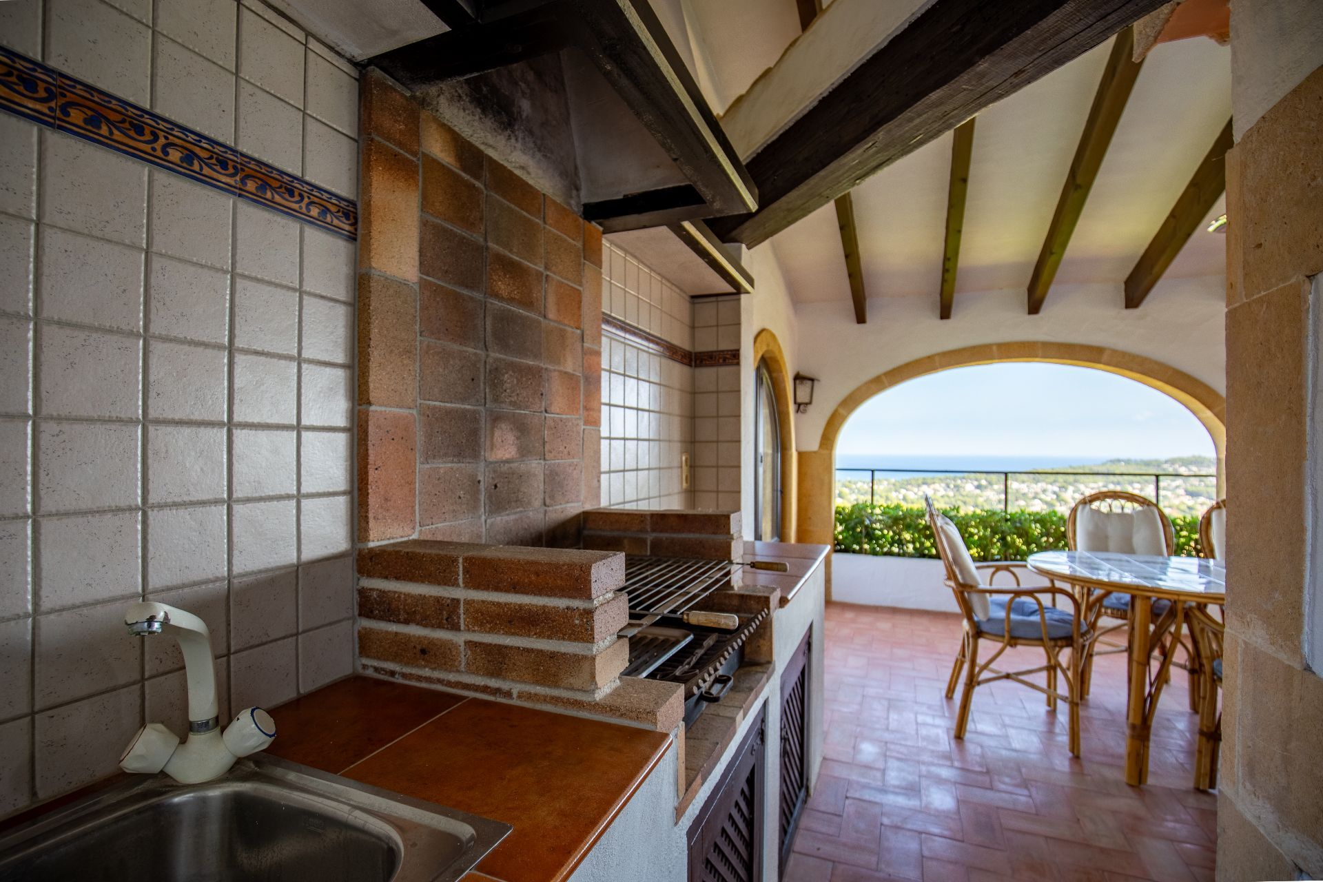 Cocina exterior cubierta con barbacoa de obra, fregadero y suelos de terracota, con vigas de madera a la vista y vistas al mar a través de un arco.