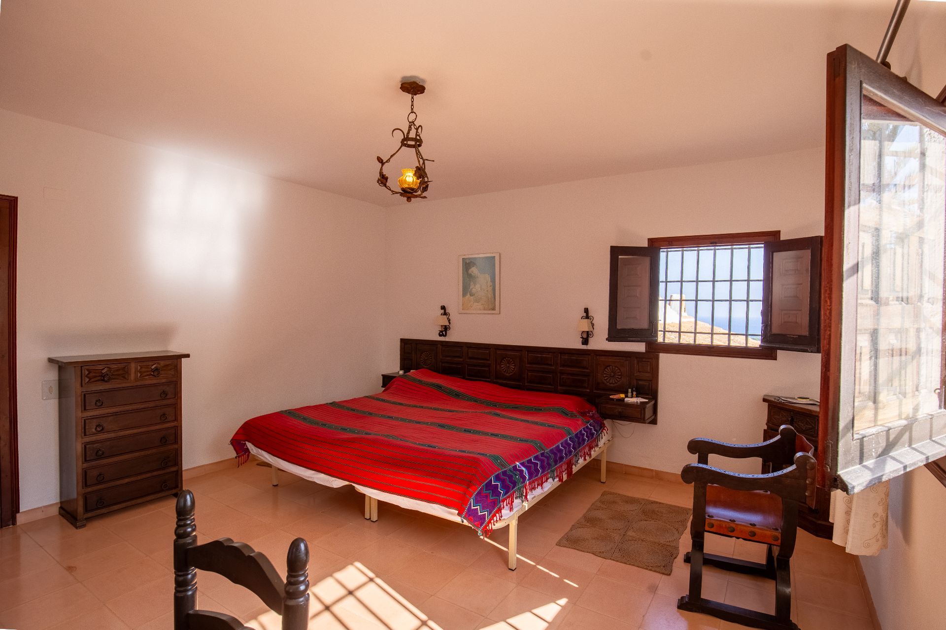 Dormitorio tradicional con cabecero de madera tallada, suelos de baldosa cerámica y lámpara de forja. La ventana con contraventanas ofrece luz natural y vistas al exterior.