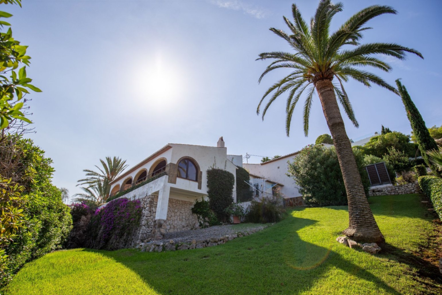 Fachada de villa mediterránea con naya de arcos, muros de piedra natural y jardín con césped inclinado y una palmera madura.