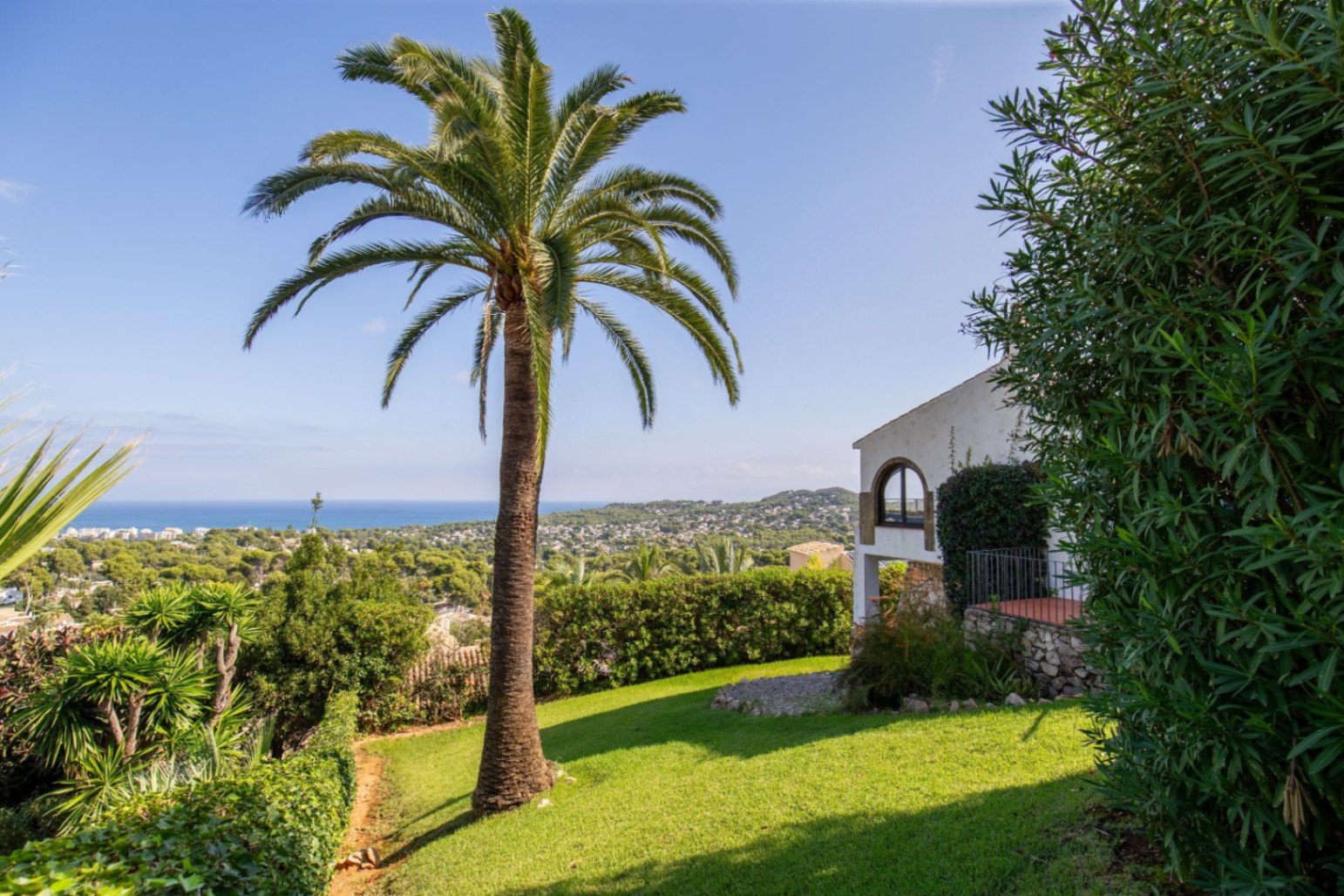 Jardín con una palmera madura y césped, con vistas a la fachada de piedra y ventana arqueada de la villa. Panorámica de la costa y el mar al fondo.