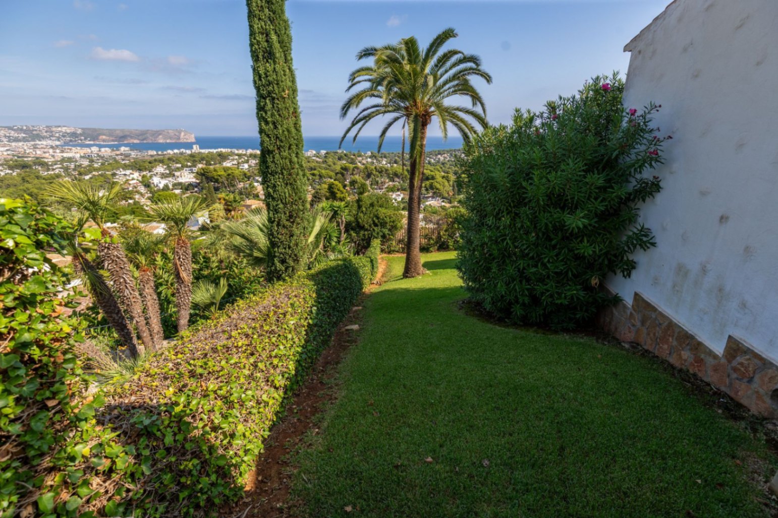 Jardín cuidado con césped, palmeras y cipreses, que ofrece vistas panorámicas de la costa y el mar Mediterráneo bajo un cielo despejado.