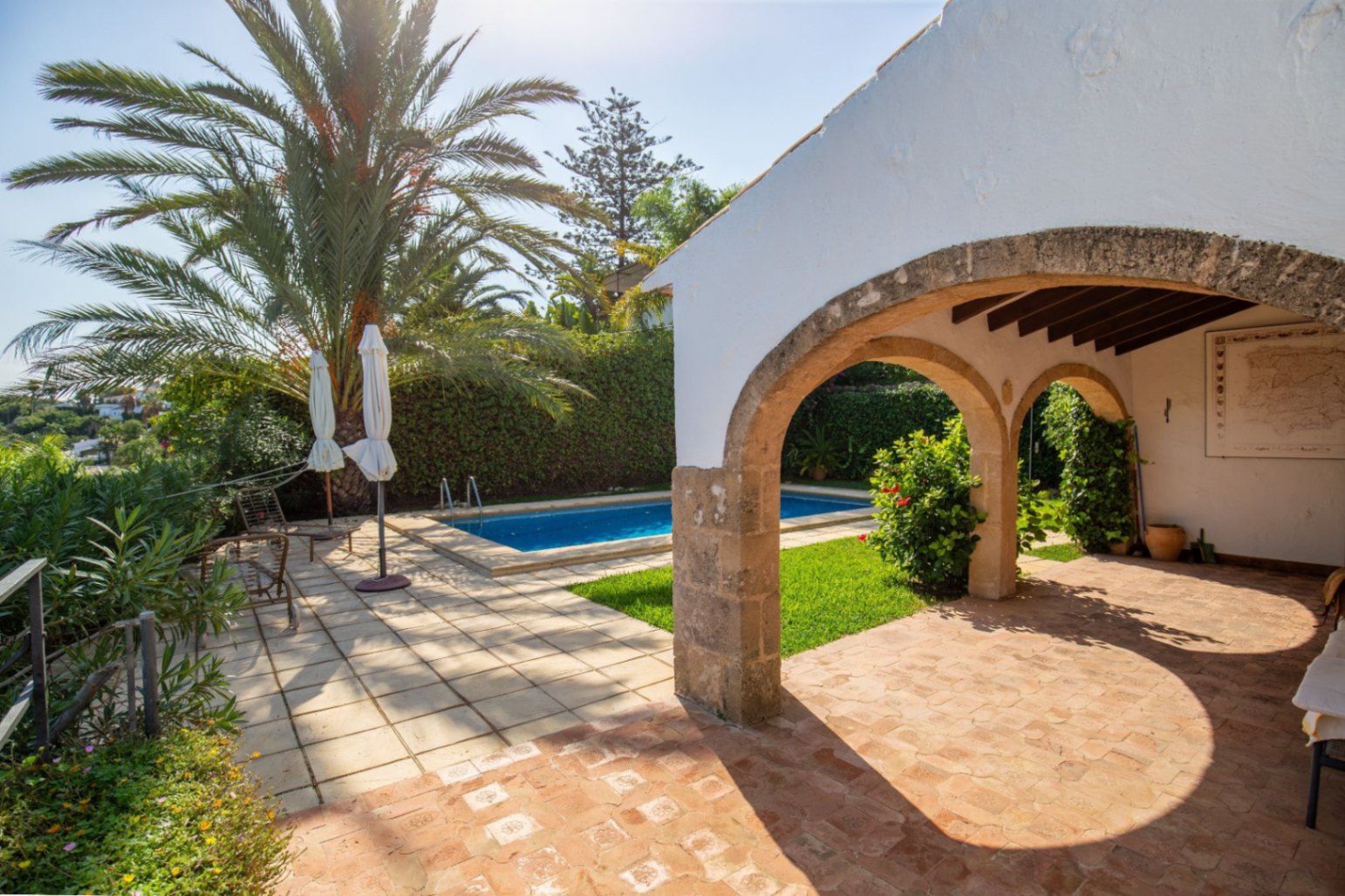 Porche con arcos de piedra natural y suelo de terracota frente a una piscina privada, rodeado de palmeras y un jardín mediterráneo consolidado.