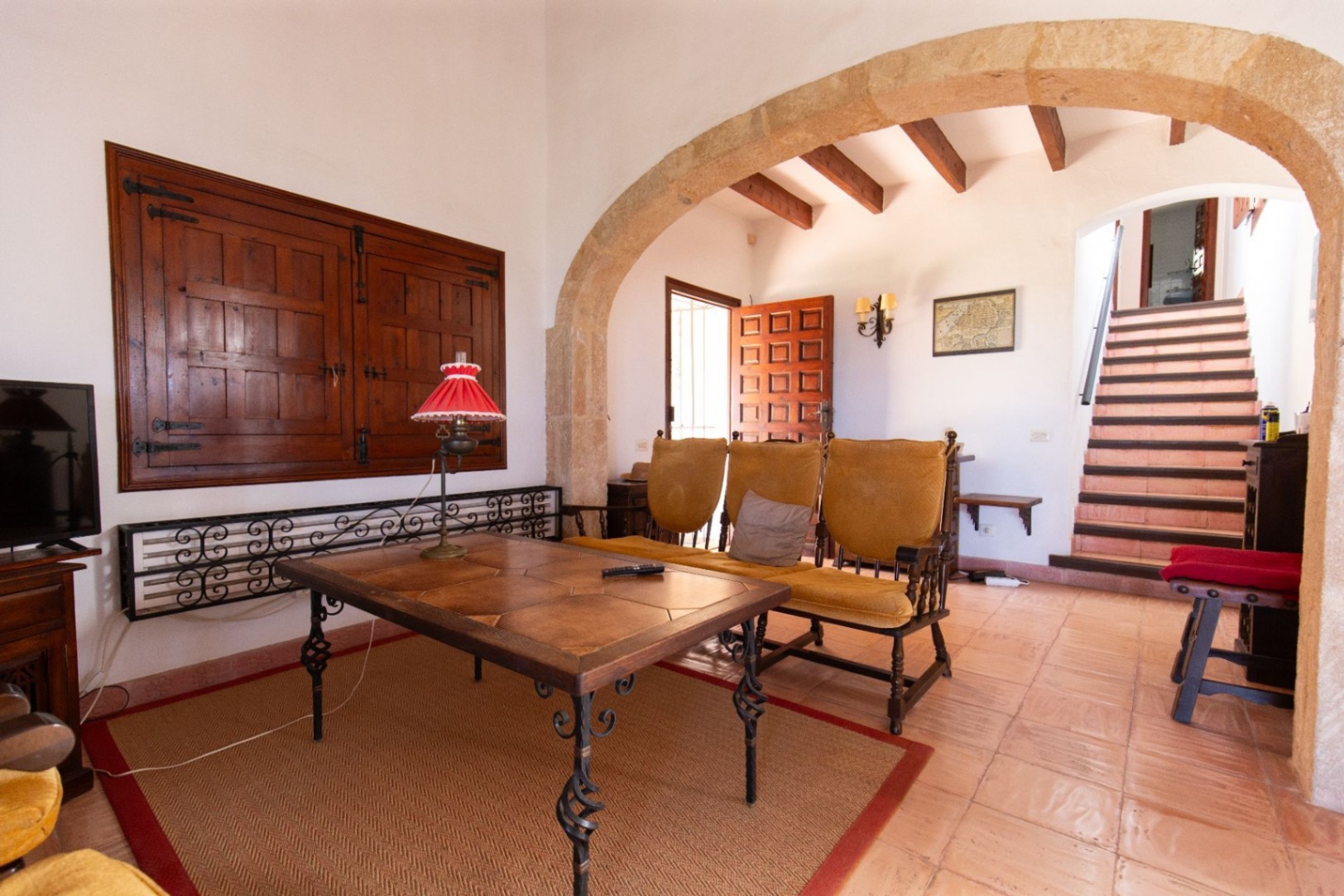 Salón con arco de piedra natural, suelos de terracota y vigas de madera vistas. Incluye mobiliario rústico y una escalera que accede a la planta superior.