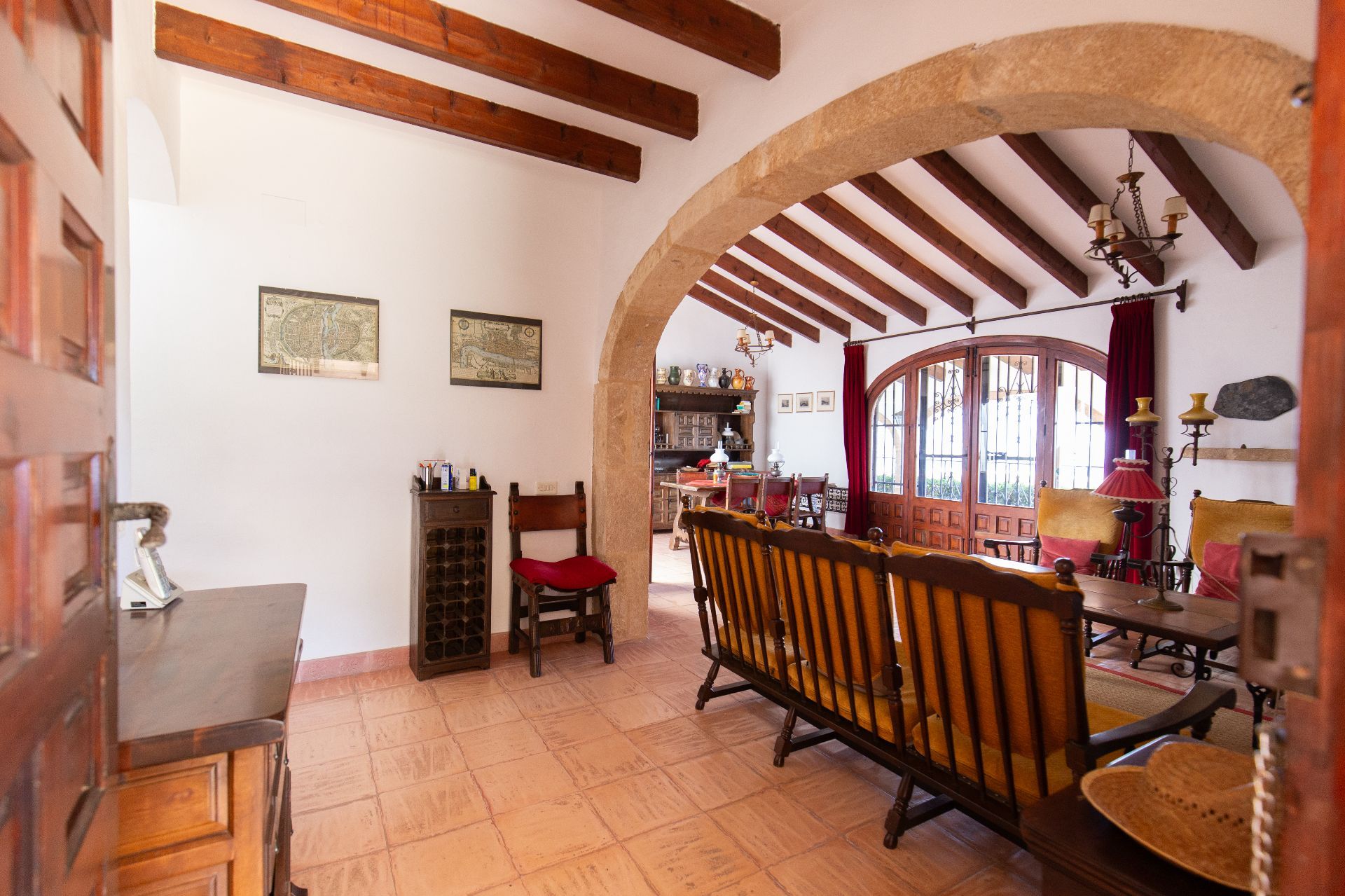 Salón rústico con vigas de madera vista, un arco de piedra natural y suelos de terracota. Incluye puertas de madera en arco con acceso al exterior y mobiliario tradicional.