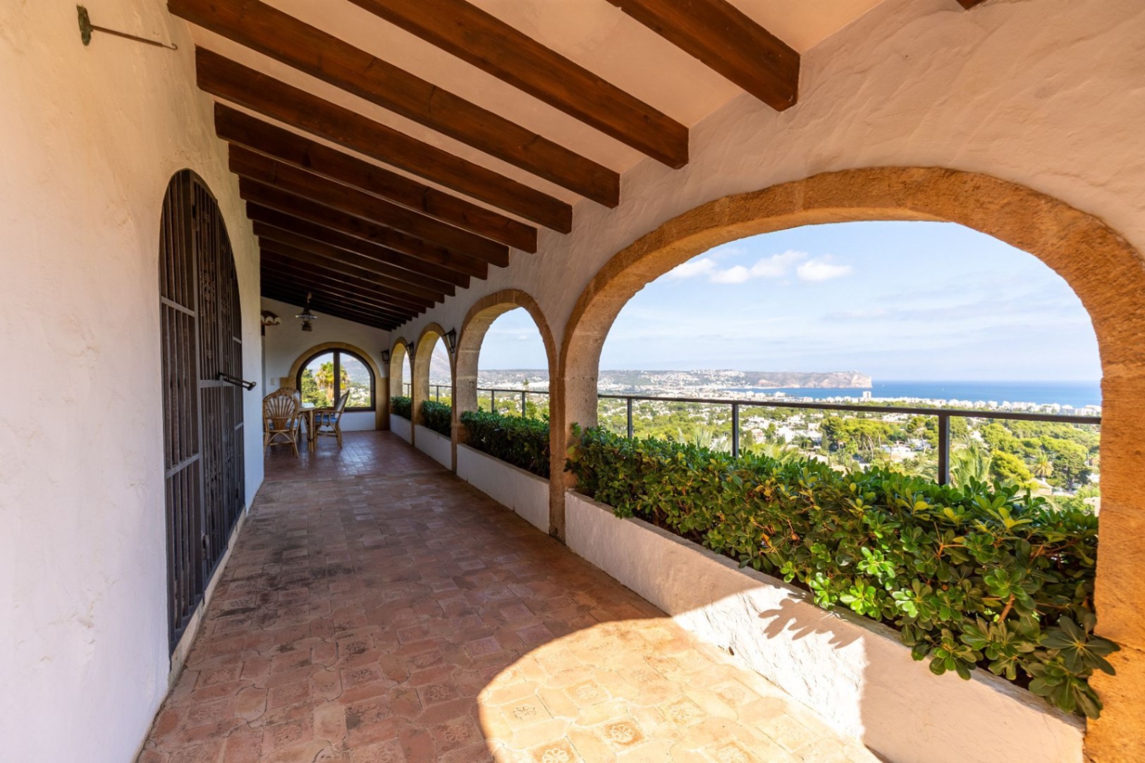 Terraza cubierta con arcos de piedra, vigas de madera a la vista y suelo de terracota. Jardineras integradas enmarcan vistas panorámicas al mar y la costa.