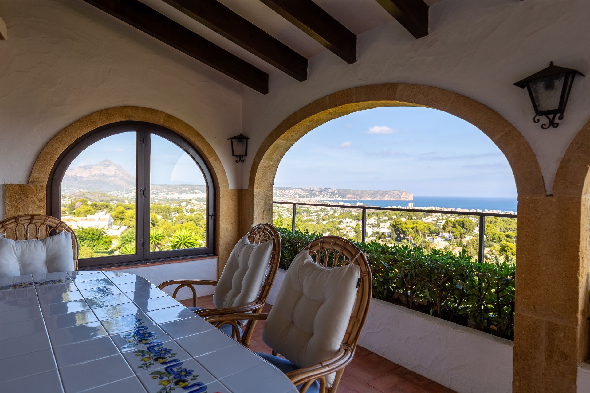 Terraza cubierta con arcos de piedra, vigas de madera y mesa de azulejos. Ofrece vistas panorámicas al mar Mediterráneo y a las montañas desde los asientos de mimbre.
