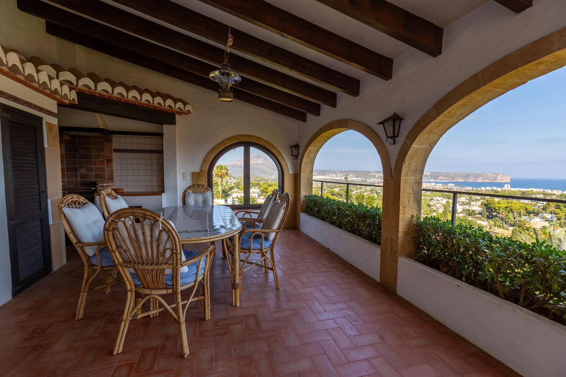 Terraza cubierta con arcos de piedra, vigas de madera y suelo de barro. Incluye comedor exterior con vistas panorámicas al mar y la montaña.