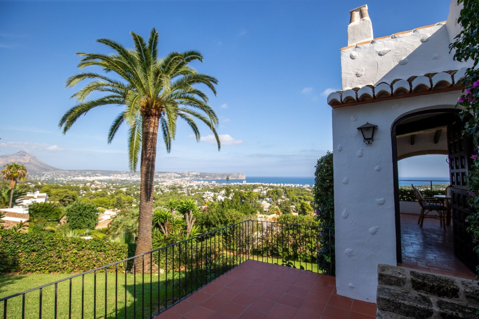Terraza de baldosas de terracota con vistas panorámicas al mar y la costa, destacando una gran palmera y la arquitectura mediterránea de paredes blancas.