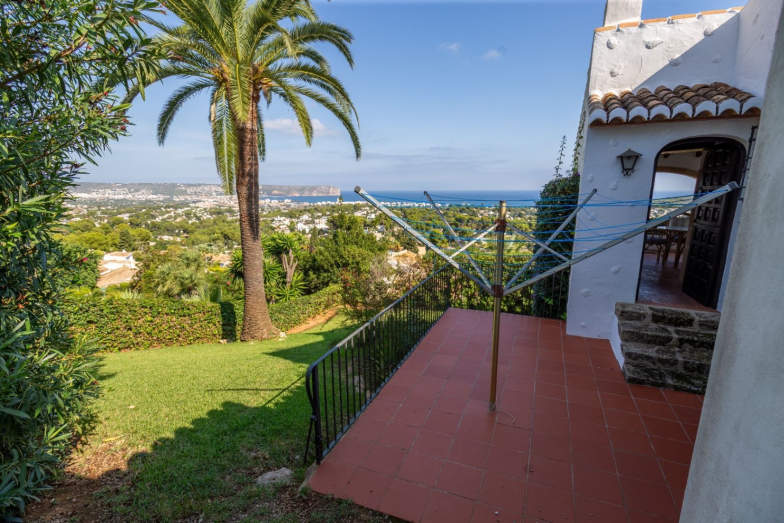 Terraza elevada con suelos de terracota y vistas panorámicas al mar y al valle costero, rodeada de un jardín con césped y una palmera madura.