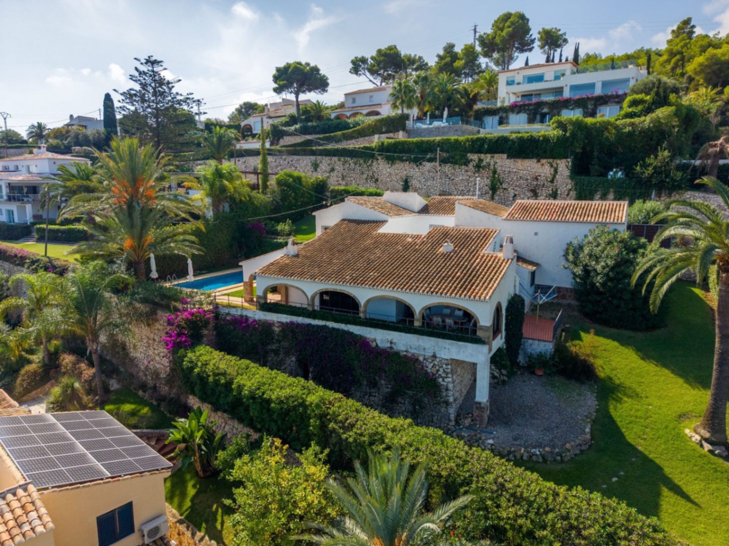 Vista elevada de una villa mediterránea con tejados de terracota, porche de arcos y piscina privada, rodeada de muros de piedra y jardines con palmeras.