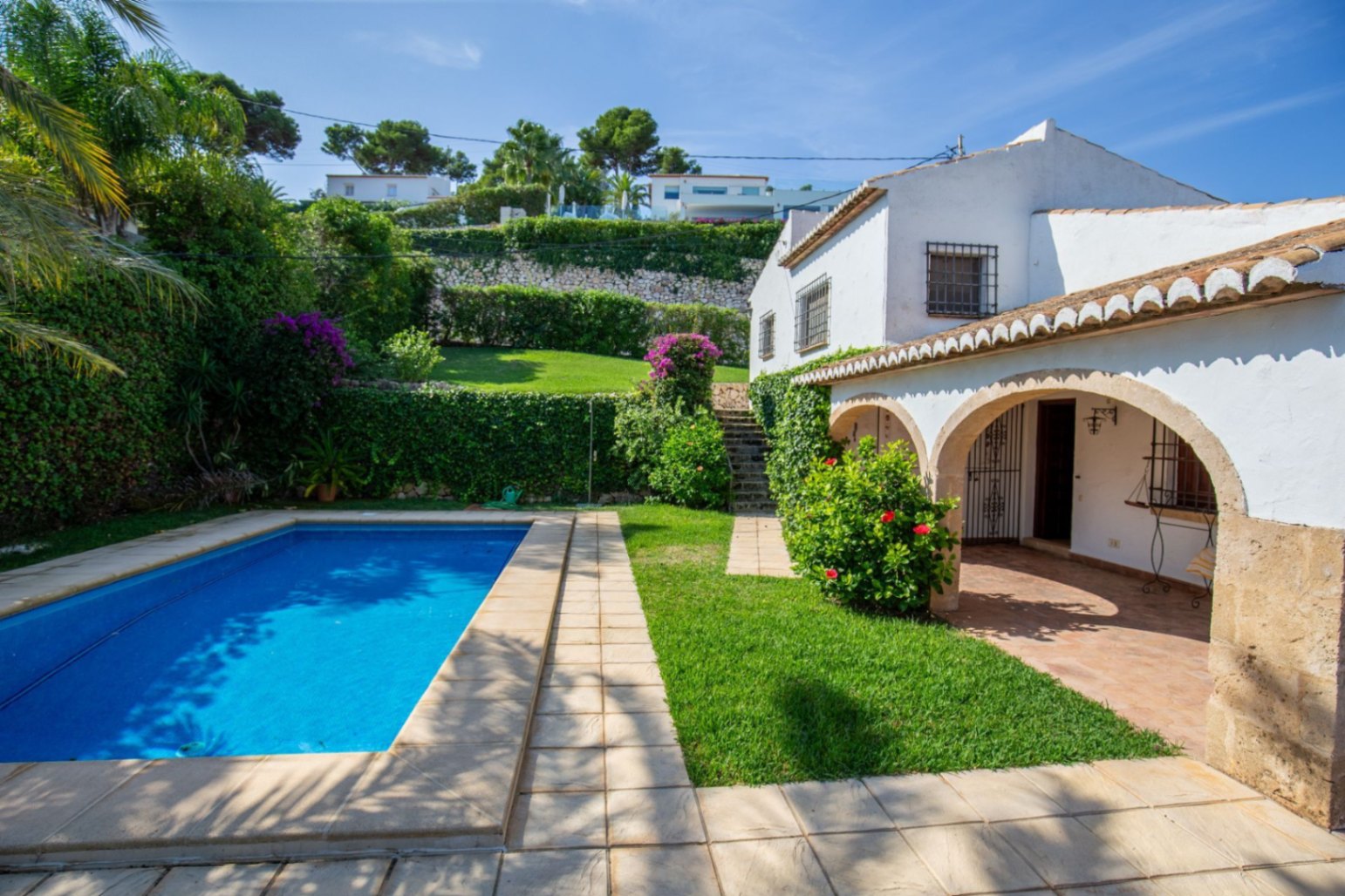 Vista exterior de una villa mediterránea con piscina rectangular, porche con arcos de piedra y jardines escalonados con buganvillas y muros de piedra.