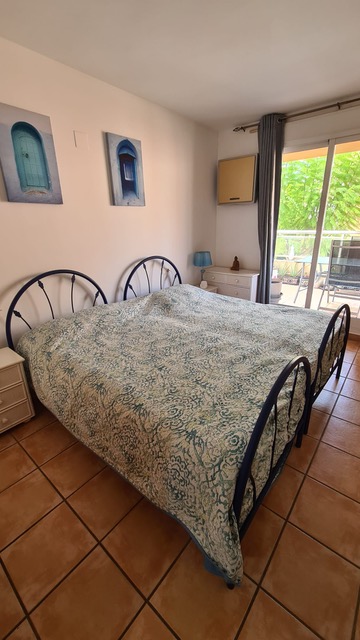 Dormitorio con cama de forja azul, suelos de gres rústico y acceso a terraza exterior mediante ventanal corredero con vistas a la vegetación.
