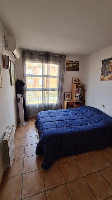 Dormitorio con suelo de gres rústico, cama con edredón azul, aire acondicionado tipo split y gran ventanal con cortinas grises translúcidas.
