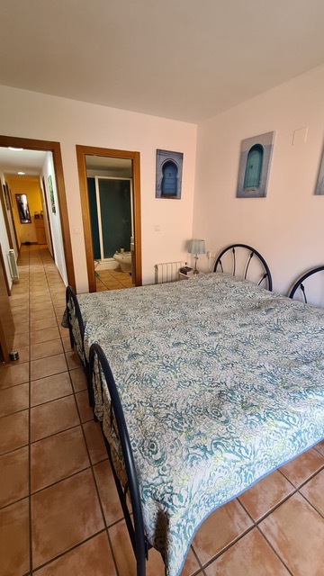 Dormitorio con suelos de gres rústico, cama con cabecero de forja y vistas hacia el baño en suite y el pasillo distribuidor de la vivienda.