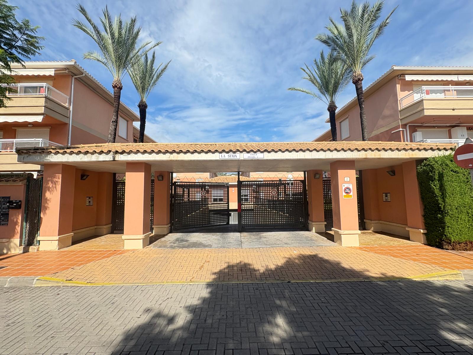 Entrada principal del complejo residencial La Senia con pilares de color terracota, techado de tejas y puertas metálicas de rejilla negra flanqueadas por palmeras.