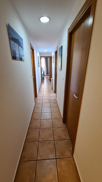 Pasillo alargado con suelos de baldosa cerámica en tonos tierra, puertas de madera natural y paredes blancas iluminadas por focos empotrados.