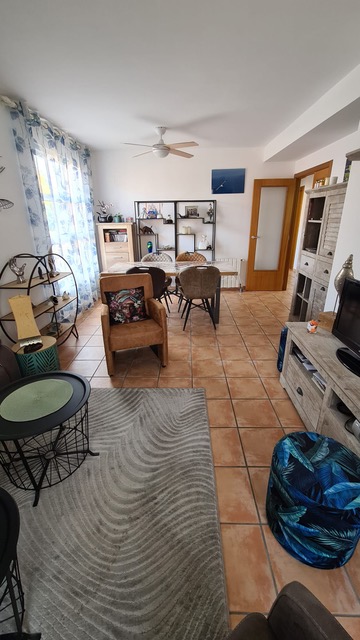 Salón comedor luminoso con suelos de baldosa de terracota, mesa de comedor de madera, ventilador de techo y zona de estar con alfombra de diseño ondulado.