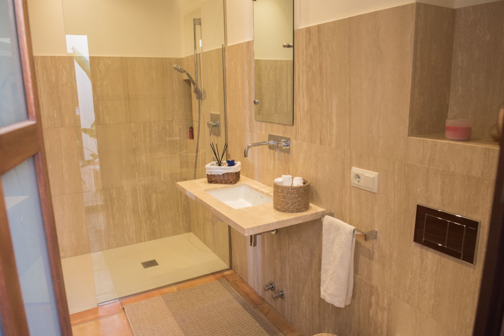 Baño moderno con ducha a ras de suelo, lavabo suspendido de piedra natural y grifería empotrada sobre revestimiento de travertino en tonos beige.