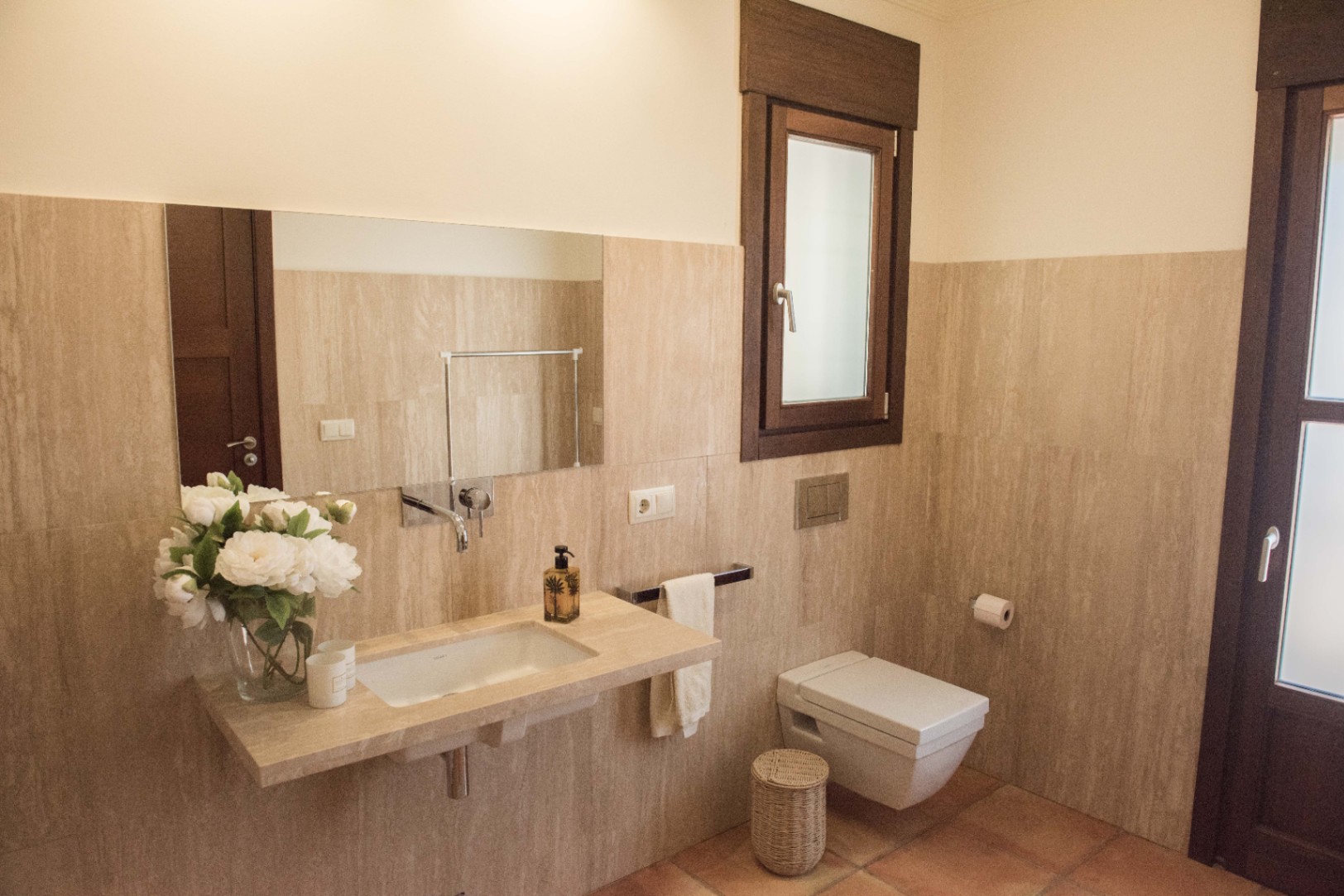 Baño moderno con lavabo integrado en encimera de piedra, grifería empotrada y sanitario suspendido sobre revestimiento de travertino y suelo de barro.