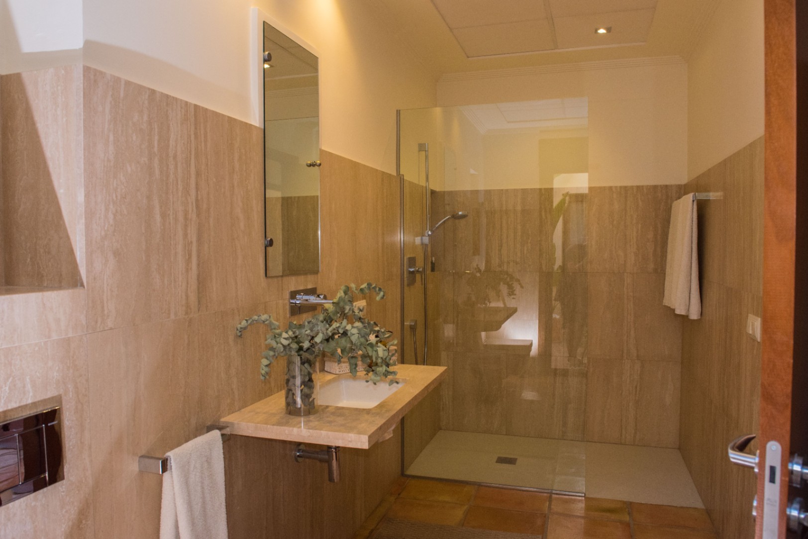 Baño moderno con revestimiento de travertino, lavabo suspendido de piedra y ducha de obra con mampara de cristal fija y suelo de terracota.