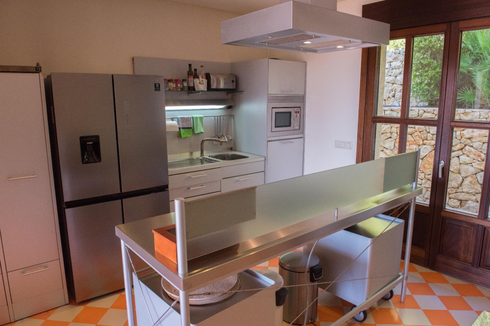 Cocina moderna con isla de acero inoxidable, suelo de gres damero y ventanales de madera con vistas a un muro de piedra seca tradicional.