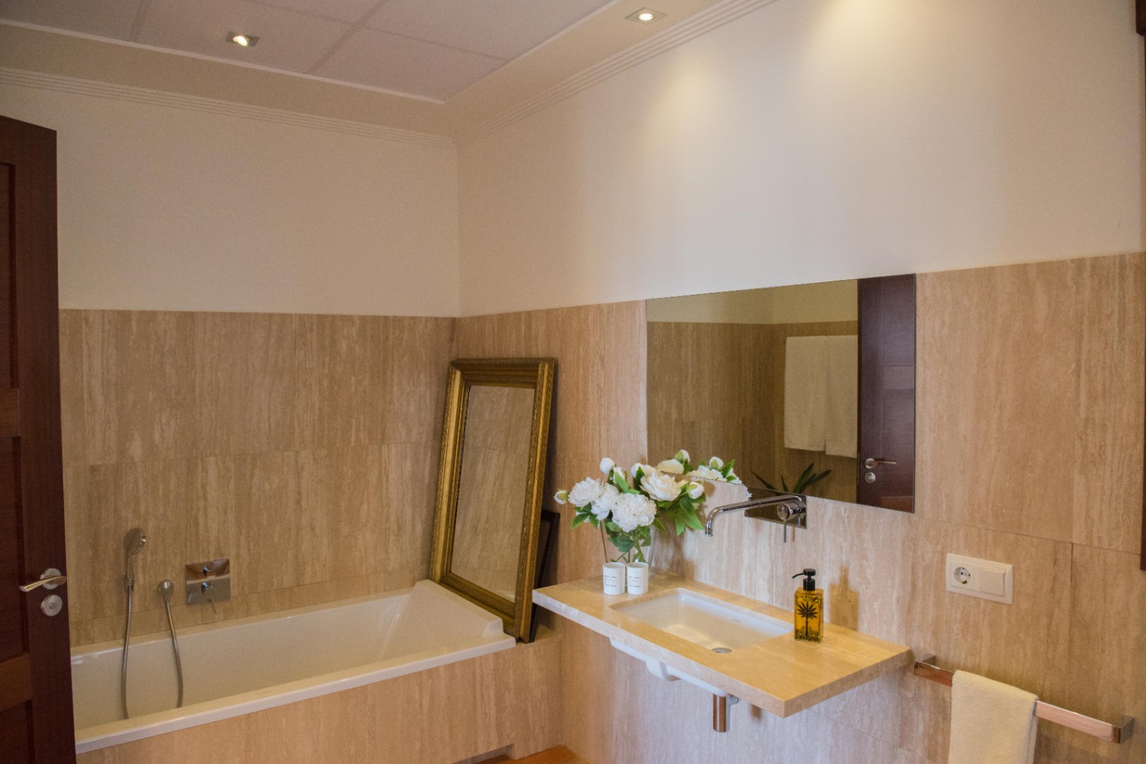 Cuarto de baño con paredes de travertino, lavabo suspendido de piedra y bañera integrada. Incluye un espejo con marco dorado y grifería cromada empotrada.