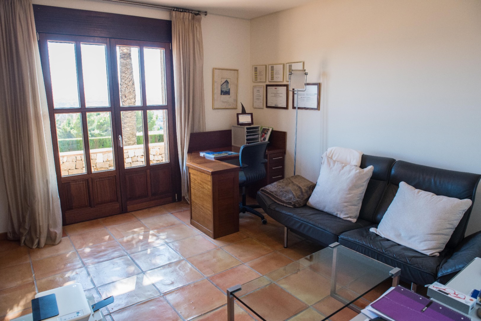 Despacho con suelos de baldosa de barro, escritorio de madera y sofá de cuero negro. Grandes puertas de madera oscura conectan con una terraza con vistas a la vegetación.