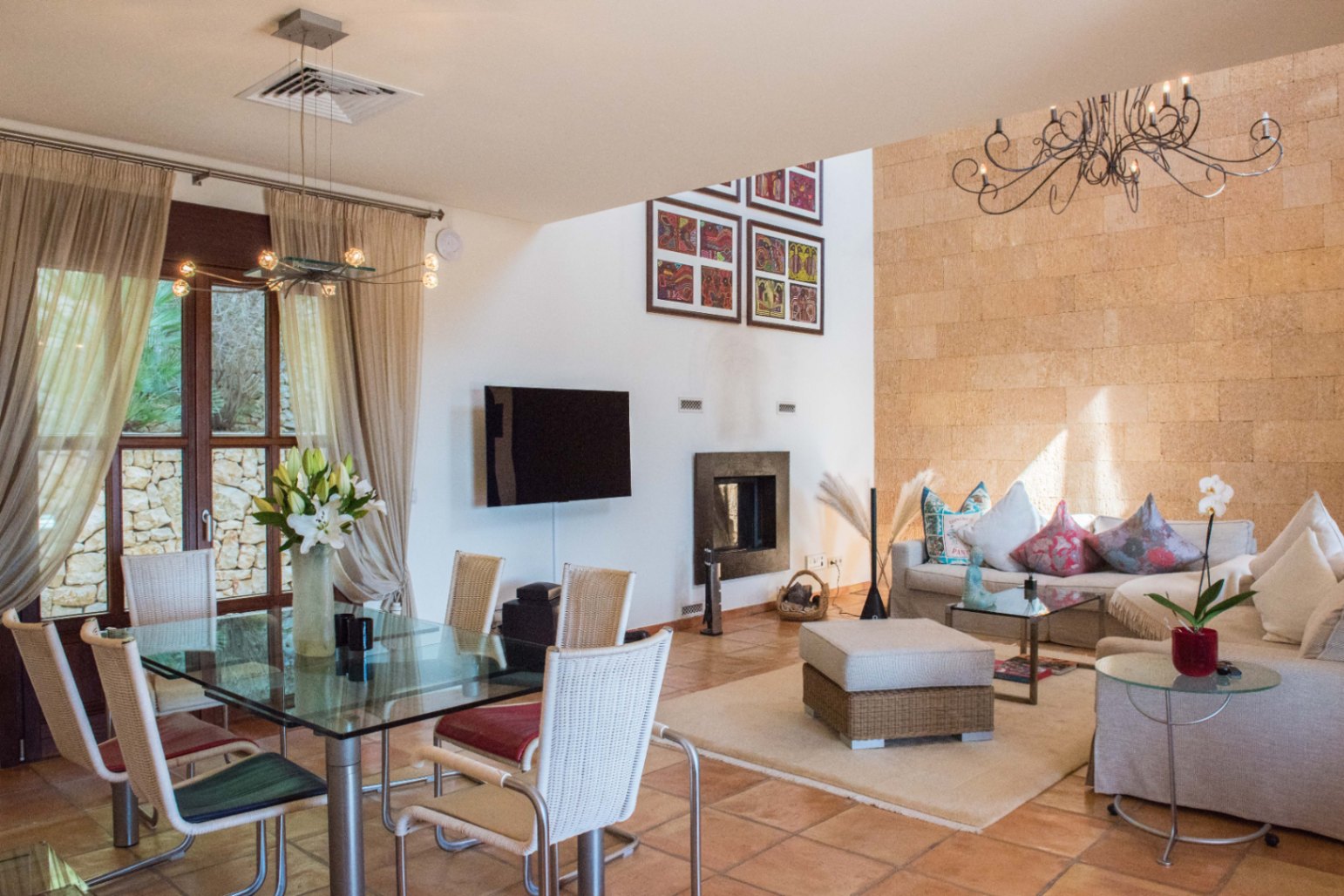 Espacio abierto con pared de acento en piedra, suelos de terracota, mesa de comedor de cristal con sillas de mimbre y zona de estar con chimenea integrada.