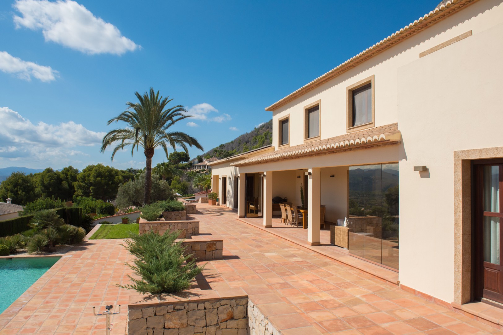 Exterior de villa mediterránea con terraza de terracota, jardineras de piedra natural y porche cubierto. Destaca la palmera junto a la piscina y las vistas despejadas.