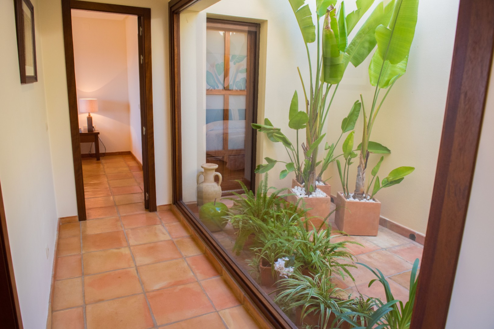 Pasillo con suelos de terracota y un gran ventanal que da a un patio interior con plantas tropicales y luz natural.