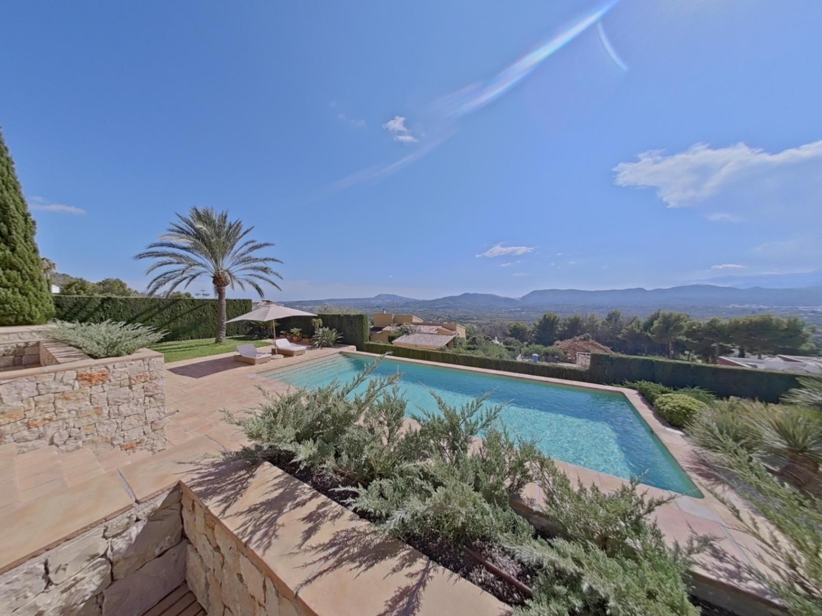 Piscina exterior con terraza de piedra y vistas panorámicas al valle. Incluye zona de tumbonas, una palmera y vegetación mediterránea cuidada.