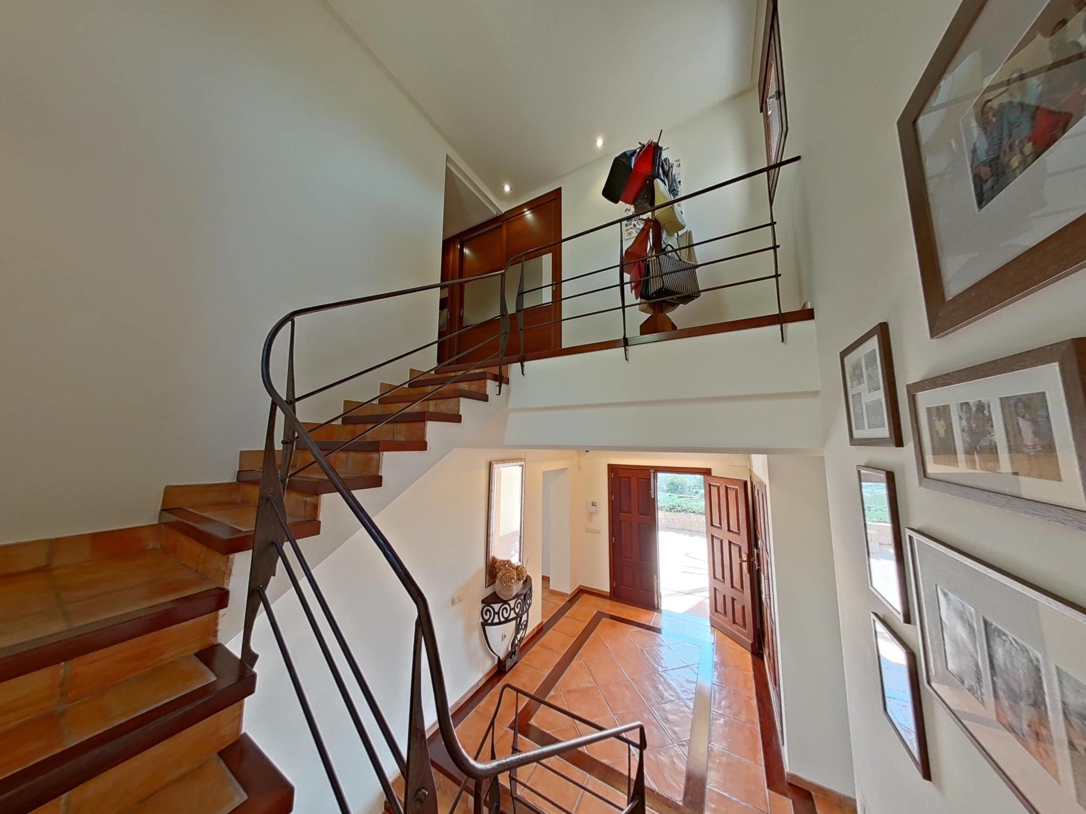 Recibidor visto desde la escalera, con suelos de baldosa de terracota, barandilla de hierro forjado y una puerta doble de madera que da al exterior.