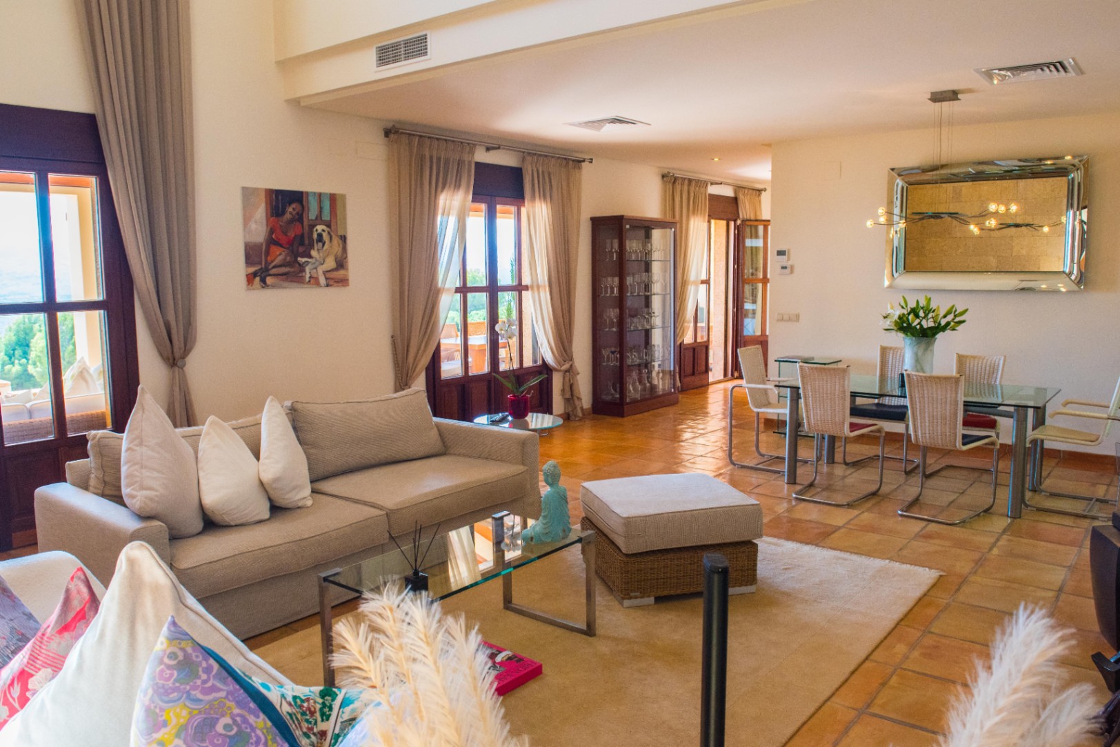 Salón comedor de planta abierta con suelos de terracota, mesa de comedor de cristal y acceso a la terraza a través de ventanales de madera con cortinas.