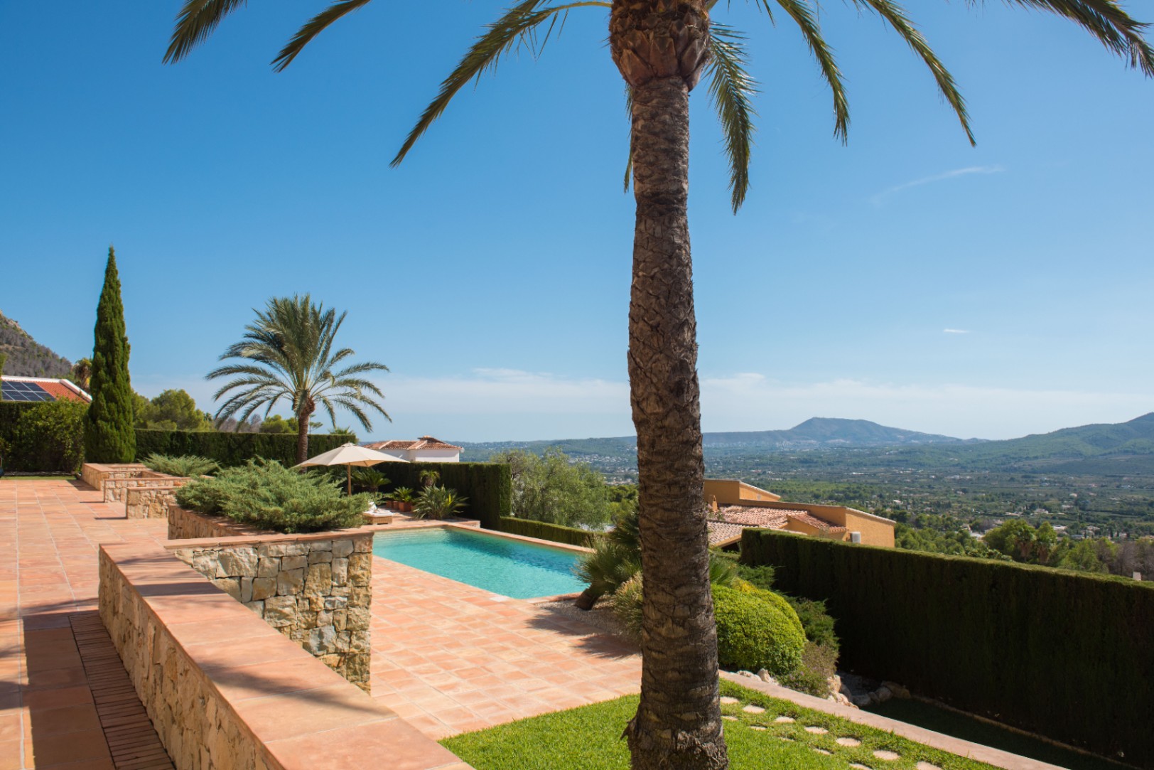 Terraza con suelos de terracota y muros de piedra natural que incluye una piscina, palmeras y vistas panorámicas al valle mediterráneo.