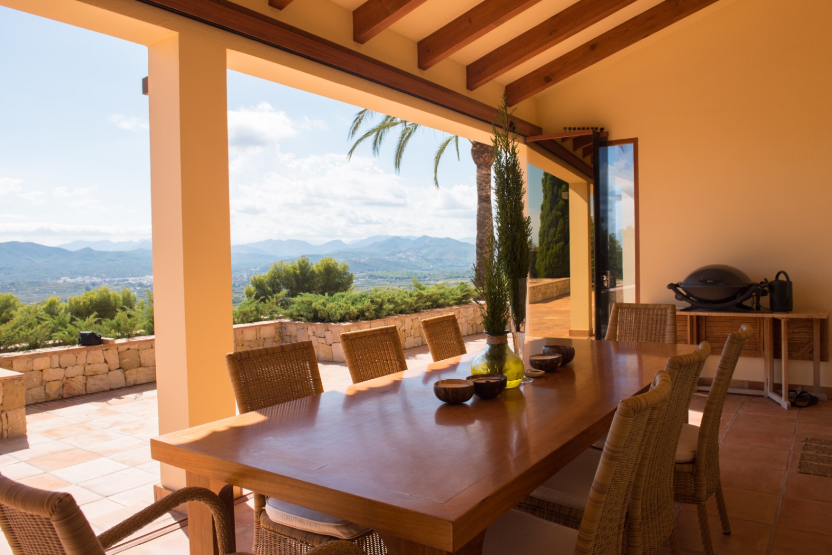 Terraza cubierta con gran mesa de madera y sillas de mimbre bajo vigas vistas. Ofrece vistas panorámicas a las montañas y suelos de terracota tradicionales.