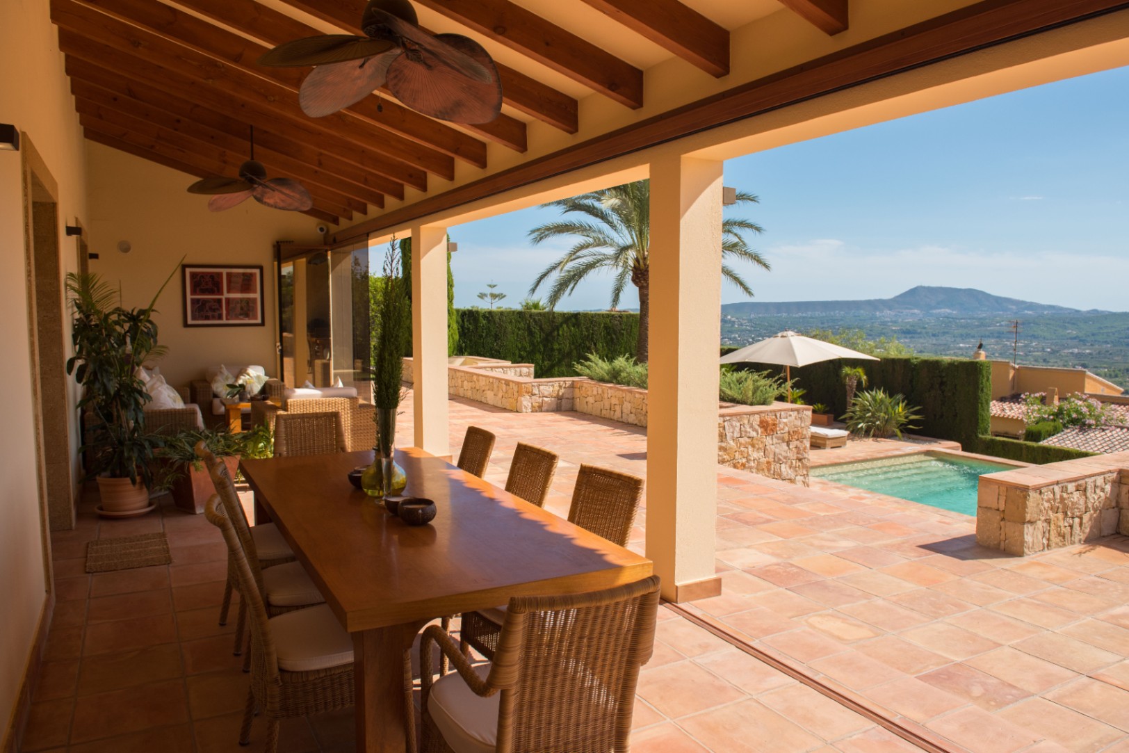 Terraza cubierta con vigas de madera, suelo de terracota y comedor exterior con vistas a la piscina y al paisaje montañoso.