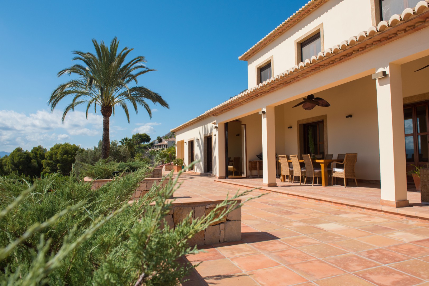 Terraza de terracota con porche cubierto, comedor exterior y vistas a una palmera y vegetación mediterránea bajo un cielo despejado.