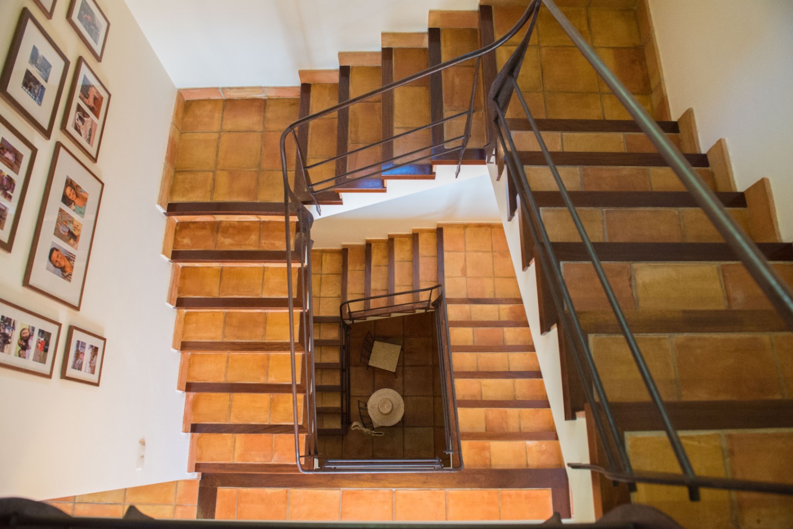 Vista cenital de una escalera de varios niveles con peldaños de terracota y bordes de madera oscura. Cuenta con una barandilla de hierro y paredes decoradas con fotografías.