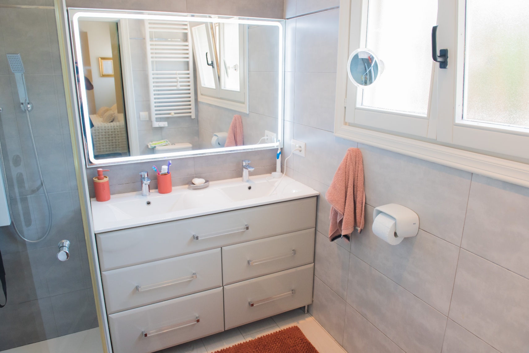 Baño moderno con mueble de doble lavabo gris, espejo retroiluminado por LED y paredes alicatadas. Dispone de ducha de obra y ventana lateral que aporta luz natural.