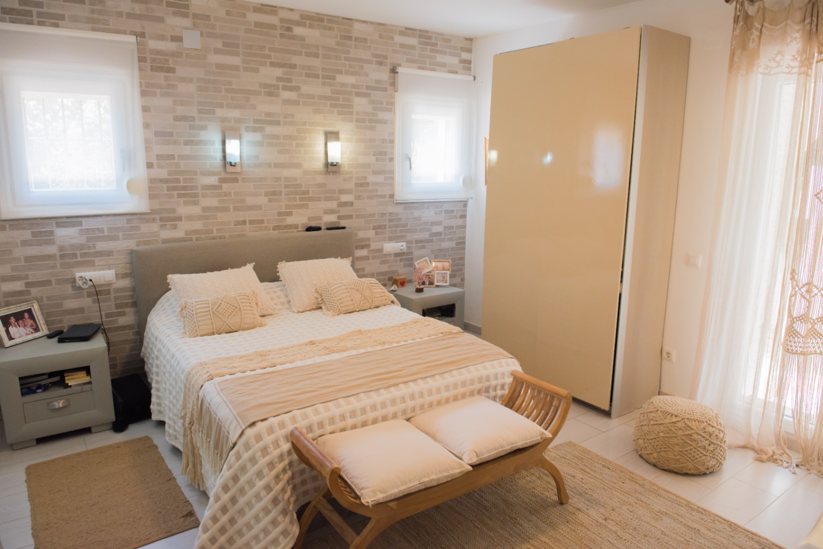 Dormitorio con pared de efecto piedra, apliques integrados, cabecero tapizado en gris y un banco de madera con cojines a los pies de la cama.