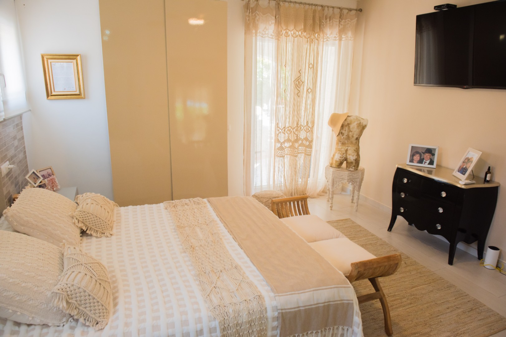 Dormitorio luminoso con cama decorada en macramé, banco de madera, armario empotrado de puertas brillantes y una cómoda negra de estilo clásico frente a la cama.