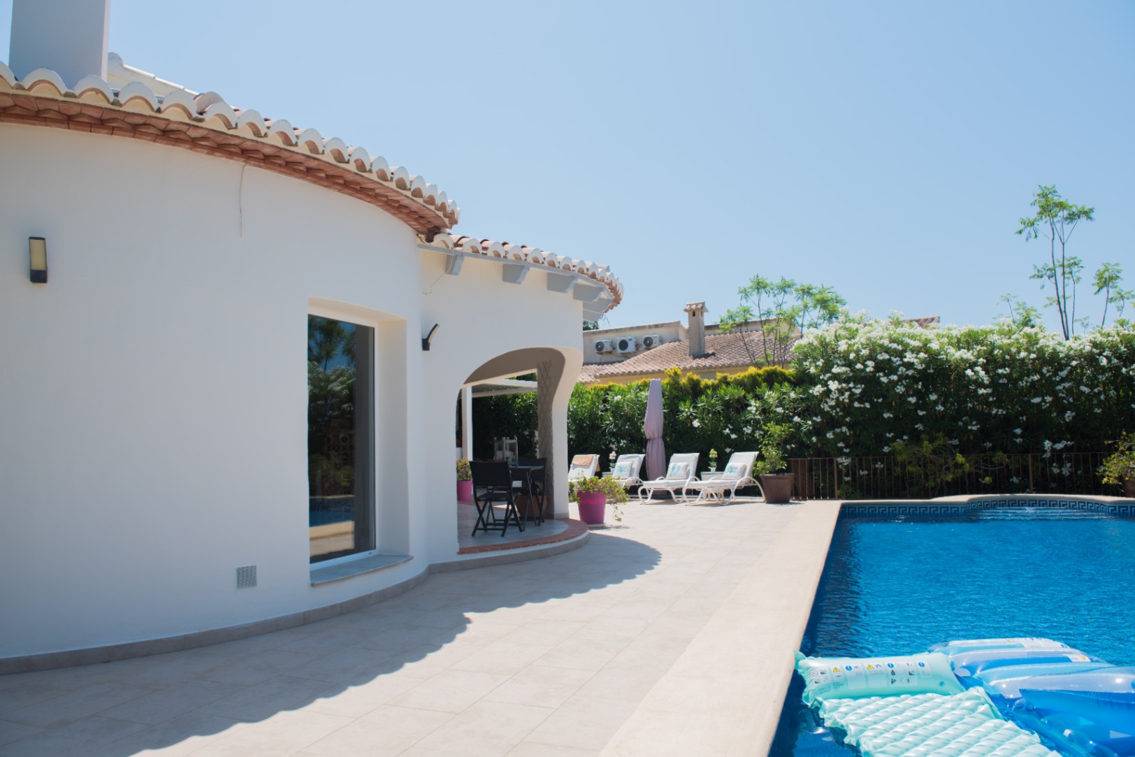 Exterior de una villa blanca con paredes curvas, terraza de piedra y piscina. El porche arqueado ofrece una zona de comedor junto a tumbonas y setos floridos.