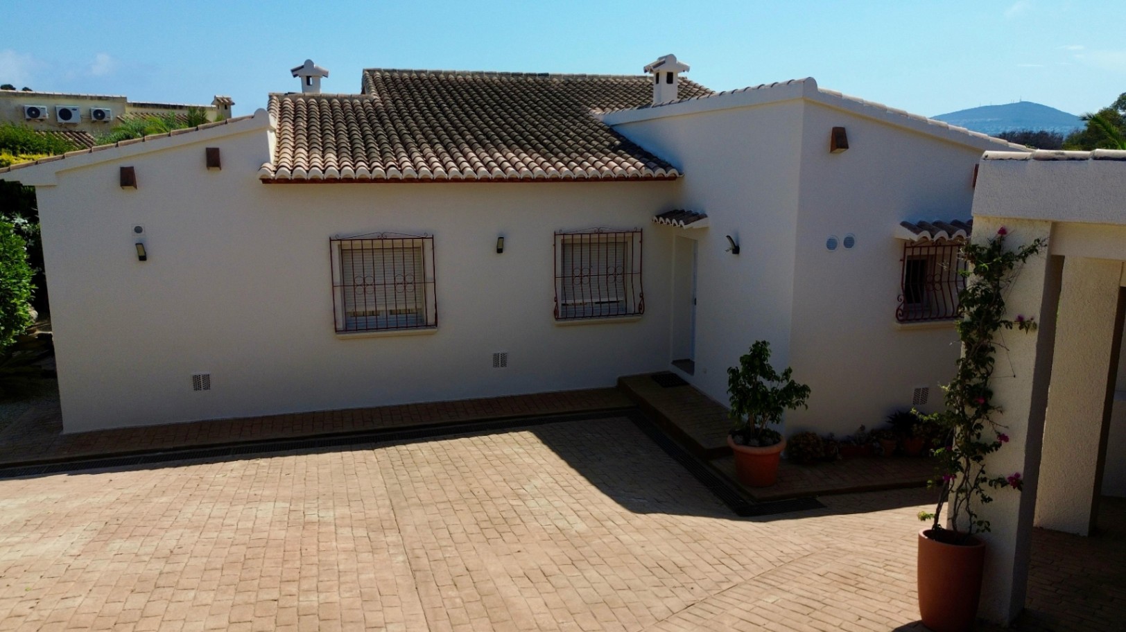 Fachada de una villa mediterránea con paredes blancas, tejado de teja cerámica y un patio pavimentado con ladrillo visto y ventanas con rejas decorativas.